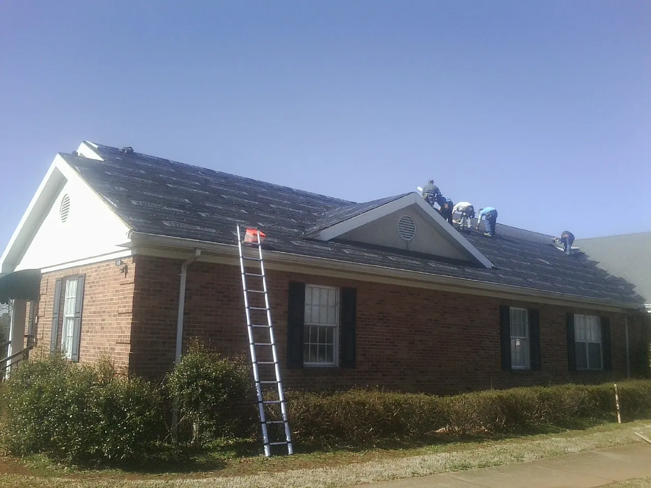 Crew tearing shingles off a brick ranch roof with a ladder set — Metal Roof Repair work by our crew in Gibsonville