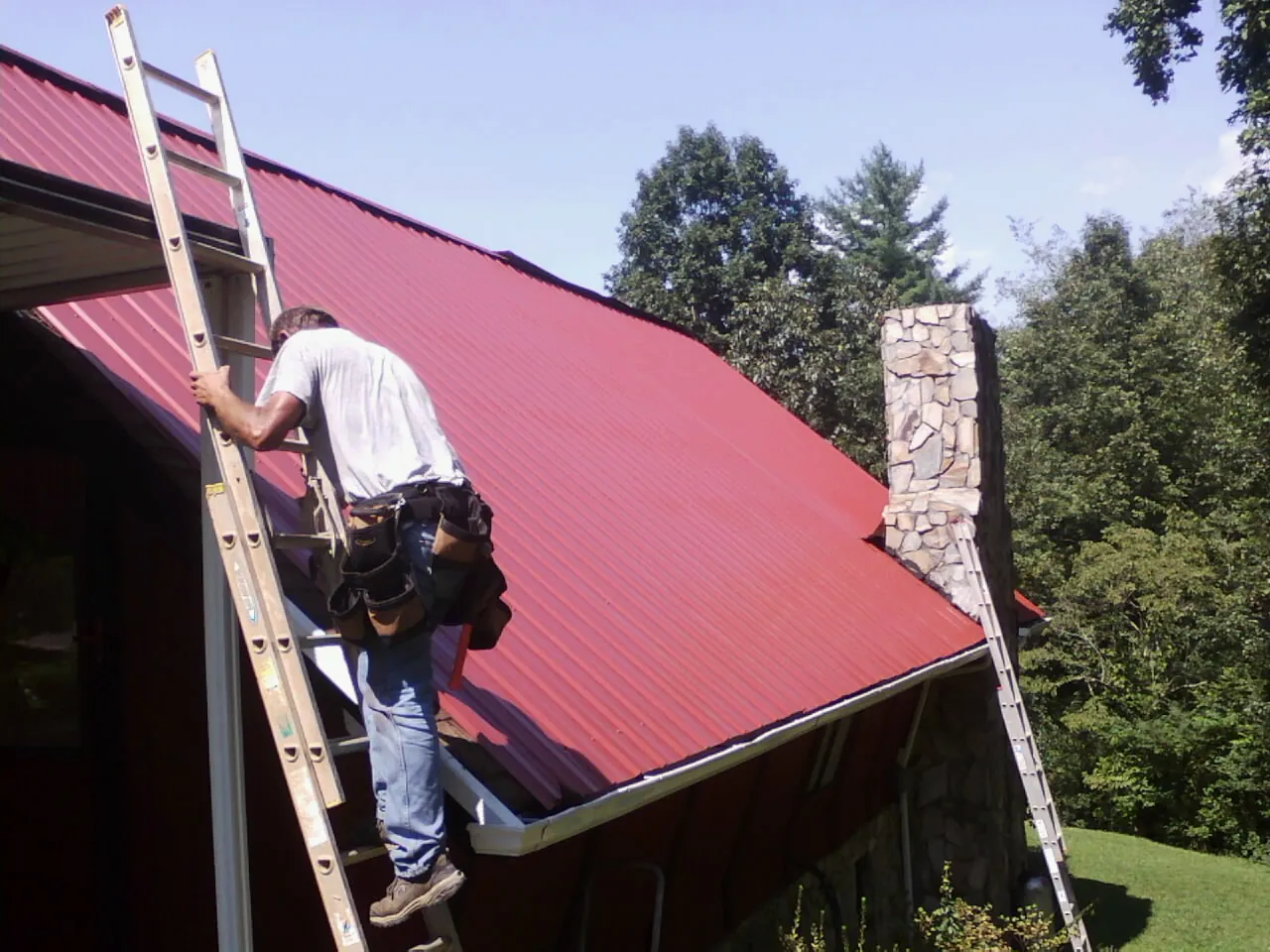 Roofer on a ladder beside a red metal roof and stone chimney — Soffit & Fascia Repair by Roofing Brothers in Gibsonville